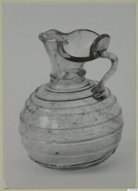 Blown glass jug with a looped handle and a wavy-rimmed spout, shown against a light grey backdrop.
