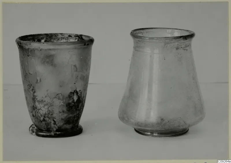 Two blown glass vessels, a beaker and a jar, standing side by side on a pale surface