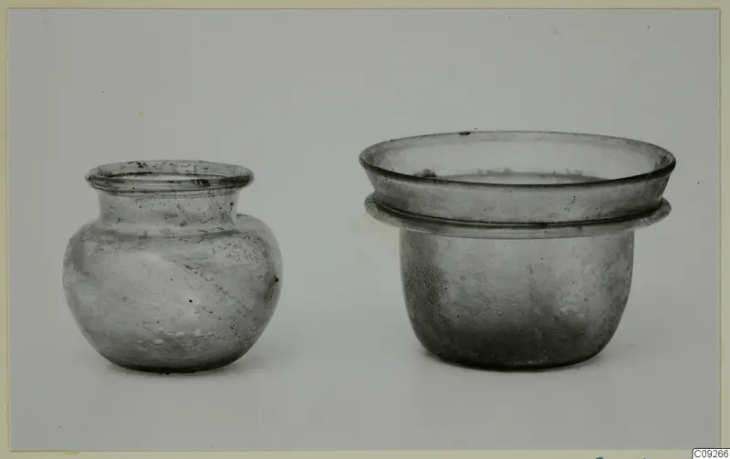 Two small glass vessels: a round-bodied jar on the left and a wide cup-like pyxis with a flanged rim on the right.