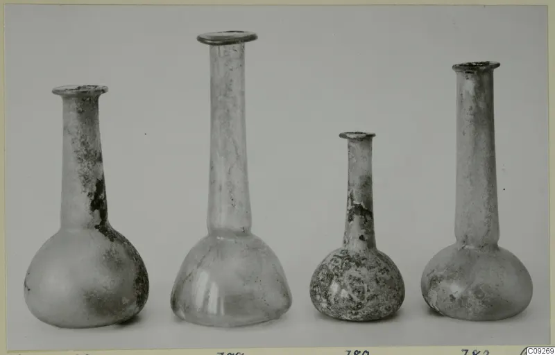 Four small blown glass bottles with long necks standing in a row against a grey backdrop.
