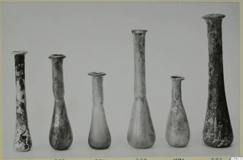 Six blown glass flasks and unguentaria of differing heights standing in a row against a grey background