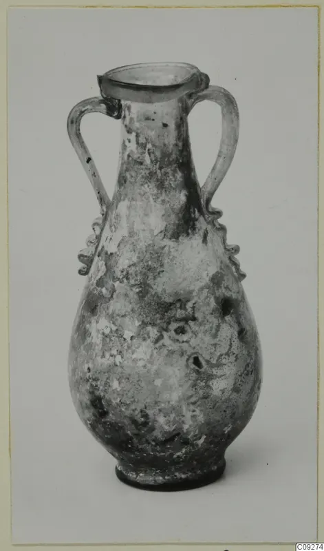 Blown glass amphoriskos with two handles and a flared rim, its surface weathered, shown against a plain grey backdrop