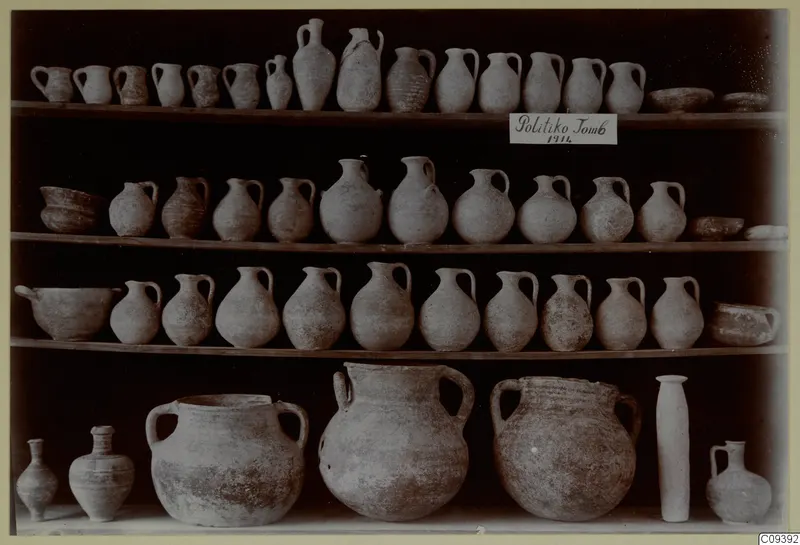 Pottery vessels from Politiko Tomb 1914 arranged on tiered wooden shelves: handled jugs, bowls and large jars.