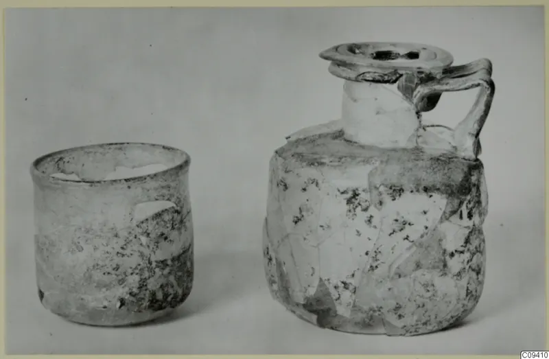 Two weathered glass vessels on a grey backdrop: a straight-sided beaker and a jug with a strap handle and disc-shaped mouth