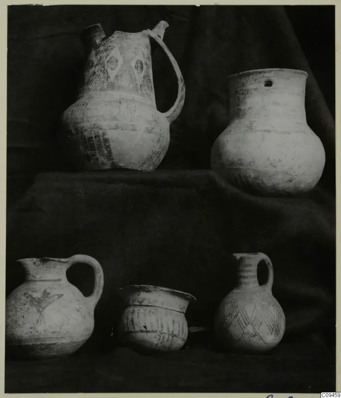 Five pottery vessels, including jugs, a jar and a bowl, arranged on dark draped cloth in two tiers