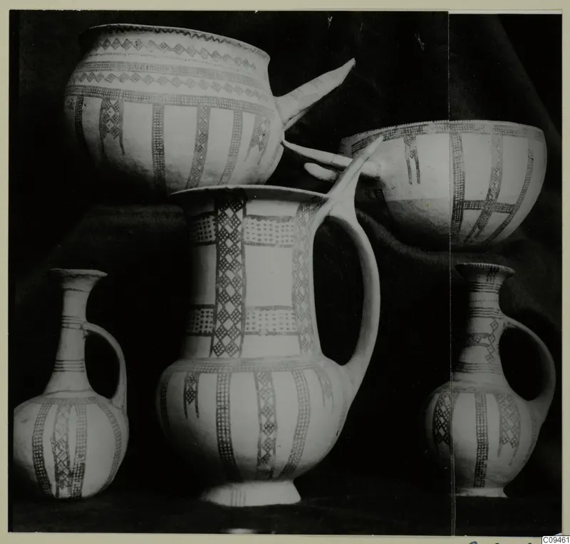 Group of painted pottery vessels: two bowls, a tall jug with handle, and two small jugs with handles, on a dark cloth.