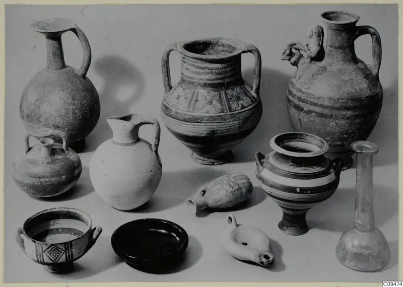 Group of about a dozen pottery vessels, a glass bottle and an oil lamp on a pale surface