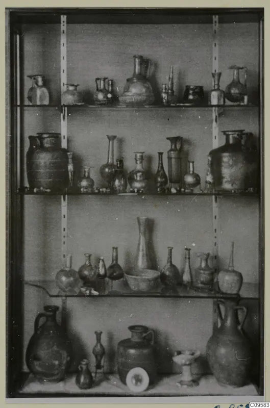 Glass vessels of many shapes displayed on four shelves in a glass-fronted case.