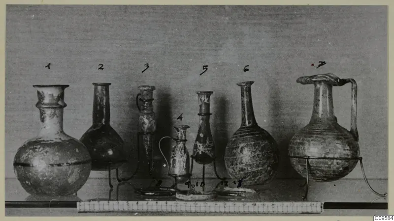 Seven blown glass vessels in a row on wire stands, numbered, with a scale bar along the front of the shelf.