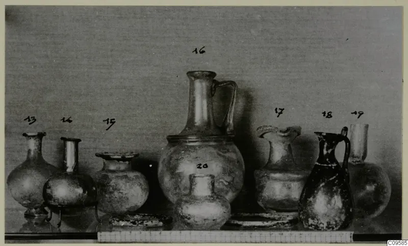 Group of eight blown glass jugs, jars and vases with numbered labels, photographed against a grey backdrop.