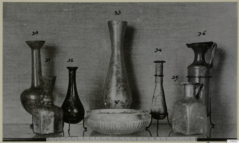 Row of blown glass jugs, vases and a ribbed bowl, each vessel with a handwritten number label above or beside it.