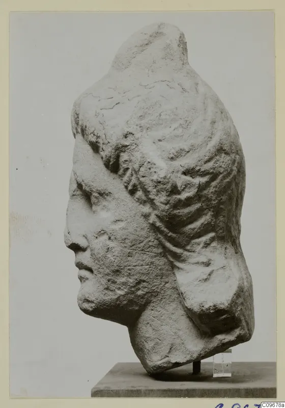Carved stone head of a figure with hair swept back in ridged waves, shown in profile on a stand.