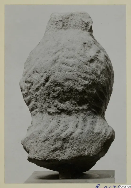 Worn stone sculpture fragment on a small dark stand, with a narrow top and a bulging lower body.