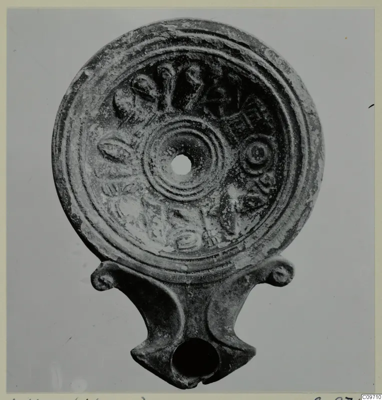 Terracotta oil lamp seen from above, with relief leaf and ring decoration around the filling hole and a nozzle with volute lugs at the base