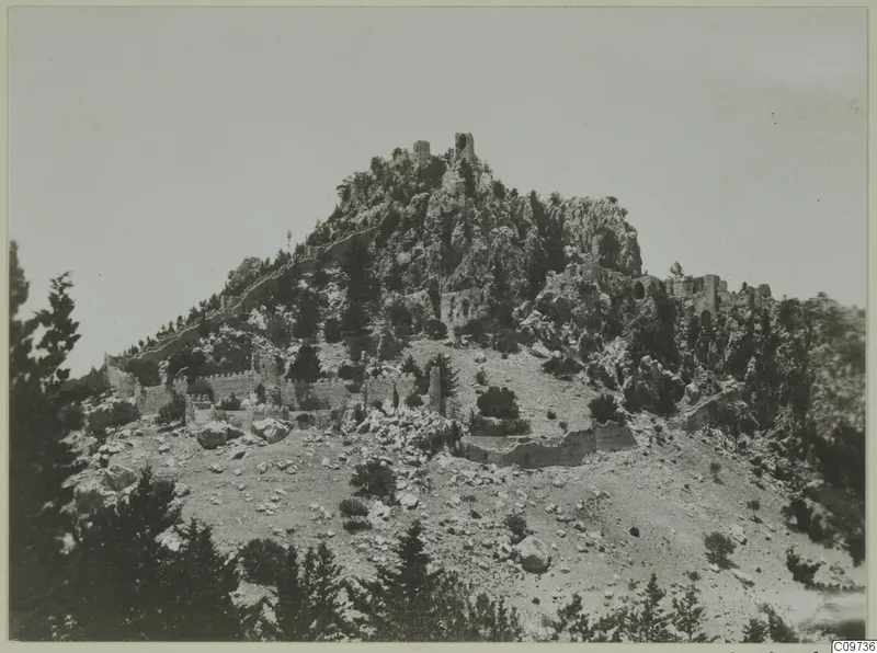Rocky hilltop with ruined walls and towers running down its slopes, with pines in the foreground.