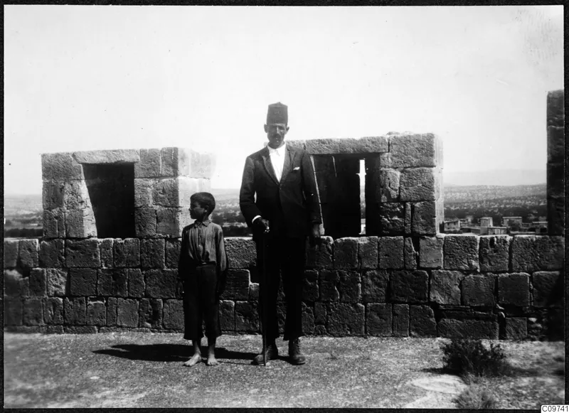 A man in a suit and fez with a cane, and a barefoot boy, on the stone walls of the fort at Kolossi