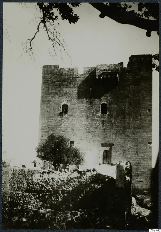 Tall square stone tower with a crenellated top, small windows and an arched doorway, with a tree in front and rubble below.