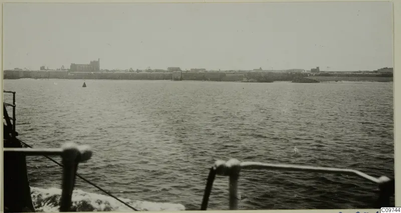 Buildings along a long sea wall, seen across calm water from the rail of a ship