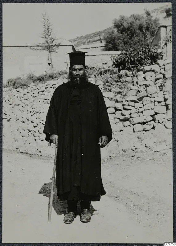 Papa Polivios standing on a dirt track in Mandres, Ayios Iakovos, holding a walking stick, with a dry-stone wall behind him.