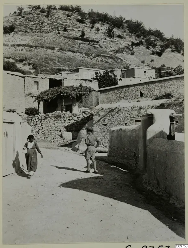 Village street at Lindros with mud-brick and stone walls, a woman walking past and a man in shorts standing in the road.