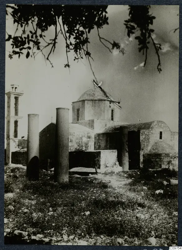 Domed building with a bell tower, two column drums in grass and a tree branch overhead, at Paphos