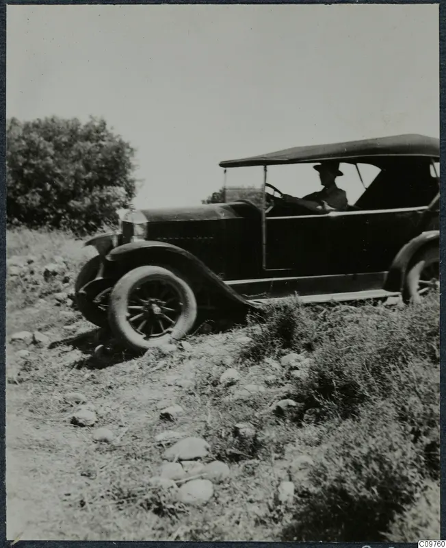 Open-topped vintage car with a driver on a stony slope beside bushes