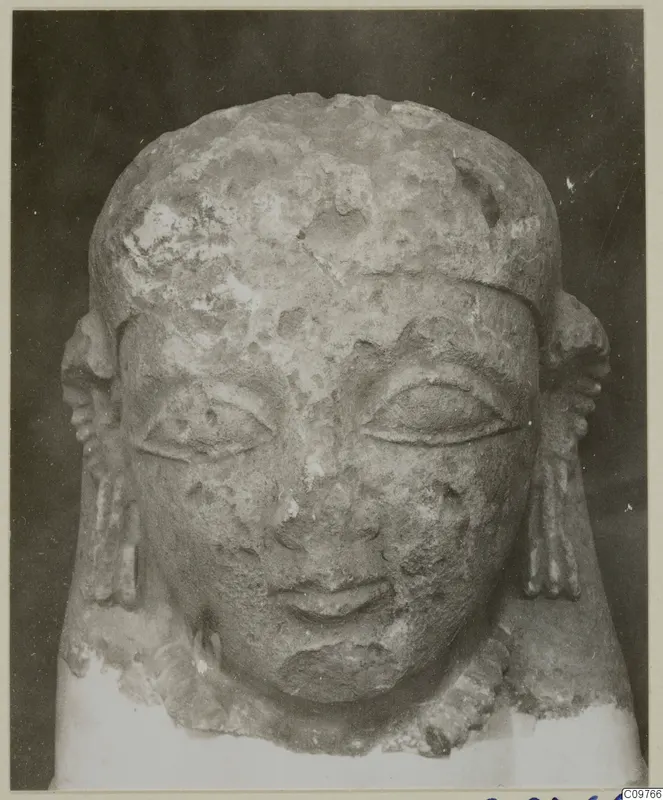 Frontal photograph of a weathered limestone sculpted head with side locks, against a dark background.