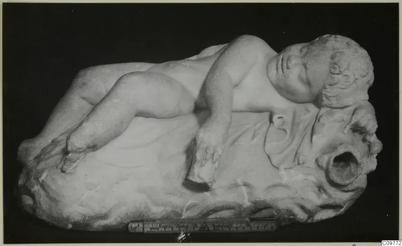 Marble sculpture of a sleeping child lying on a rocky base, with a ruler placed beside it for scale.