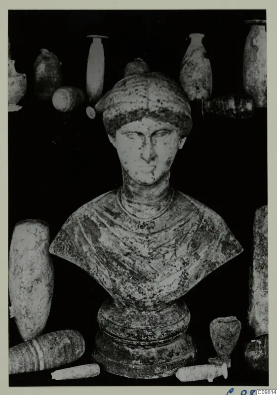 Roman sculpture: a bust of a figure with a head covering, set among several vessels on a dark background