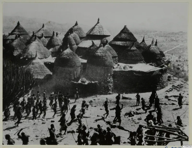 Conical thatched huts inside a walled settlement at Duvangar, with a crowd of people on bare ground in front.