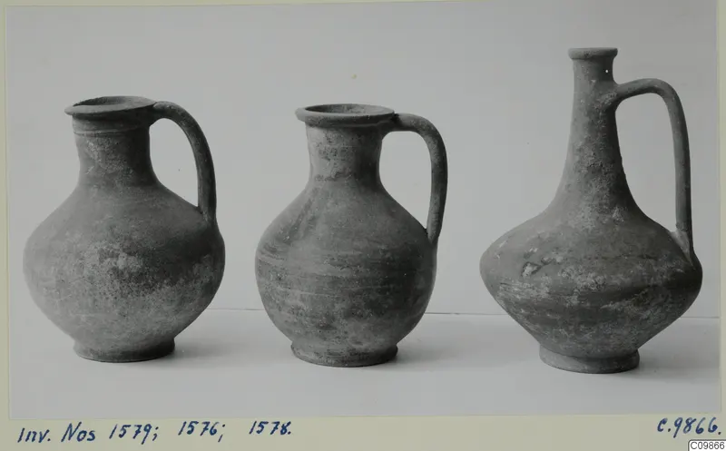 Three handled jugs of differing shapes standing in a row, photographed against a pale background.