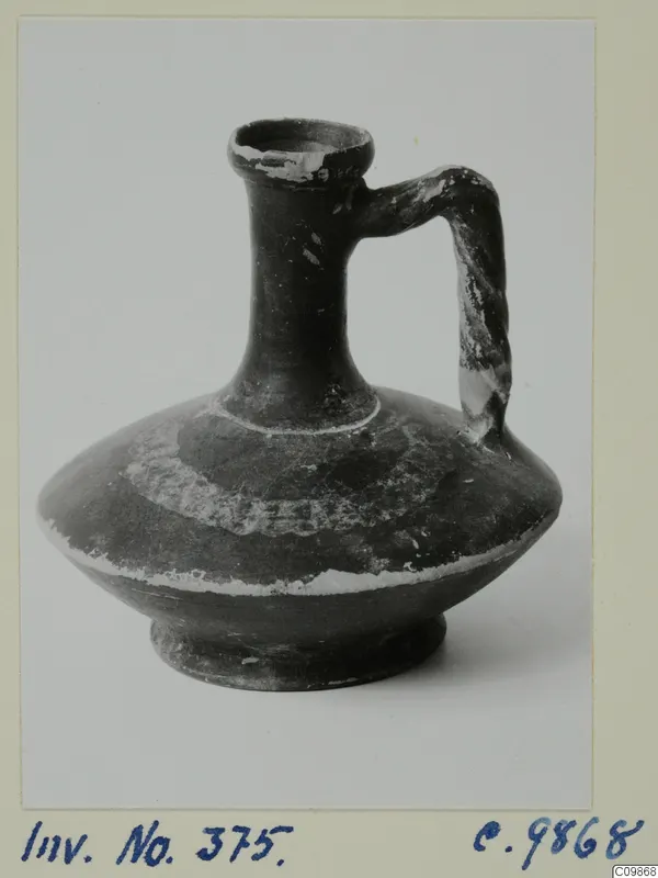 Dark jug with a tall neck, single strap handle and flaking pale paint bands on the body