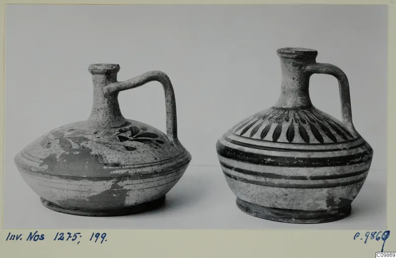 Two painted pottery jugs with single handles, side by side against a grey backdrop