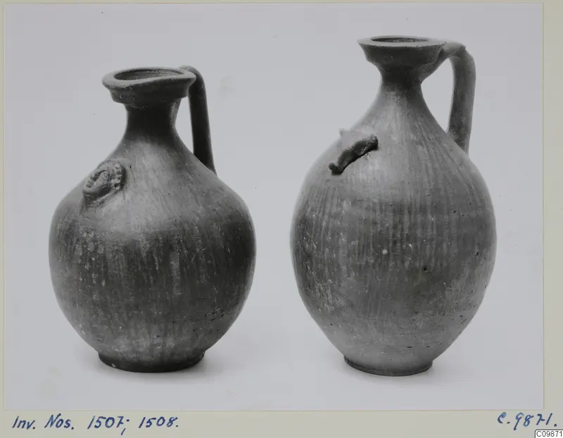 Two dark pottery jugs with single handles and applied knobs on the body, standing side by side