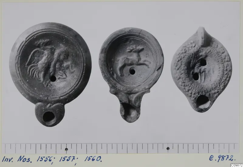 Three terracotta oil lamps with moulded decoration on the discs, shown side by side on a grey card with a scale below.