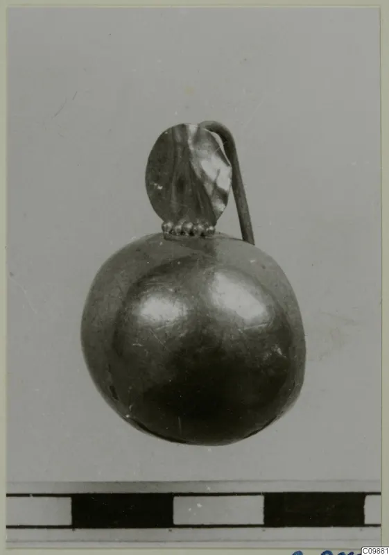 Small rounded piece of jewellery with a looped tube and a leaf-shaped plate on top, on a grey background.
