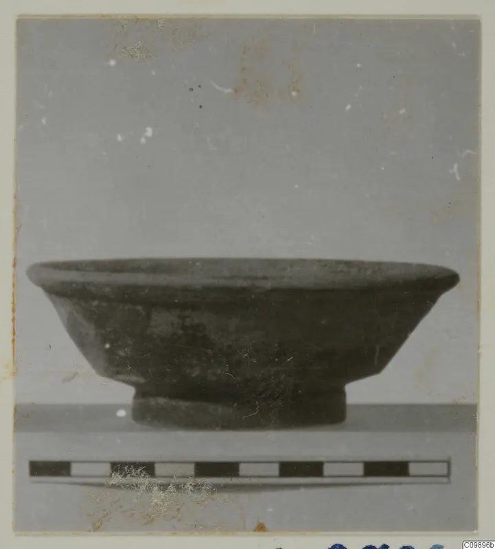 Side view of a dark pottery bowl with a flared rim and short ring foot, with a scale bar in front.