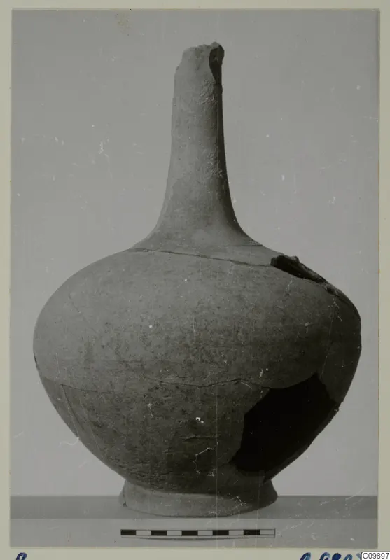 Pottery jar with a tall narrow neck, rounded body and ring foot, photographed against a grey backdrop with a scale bar