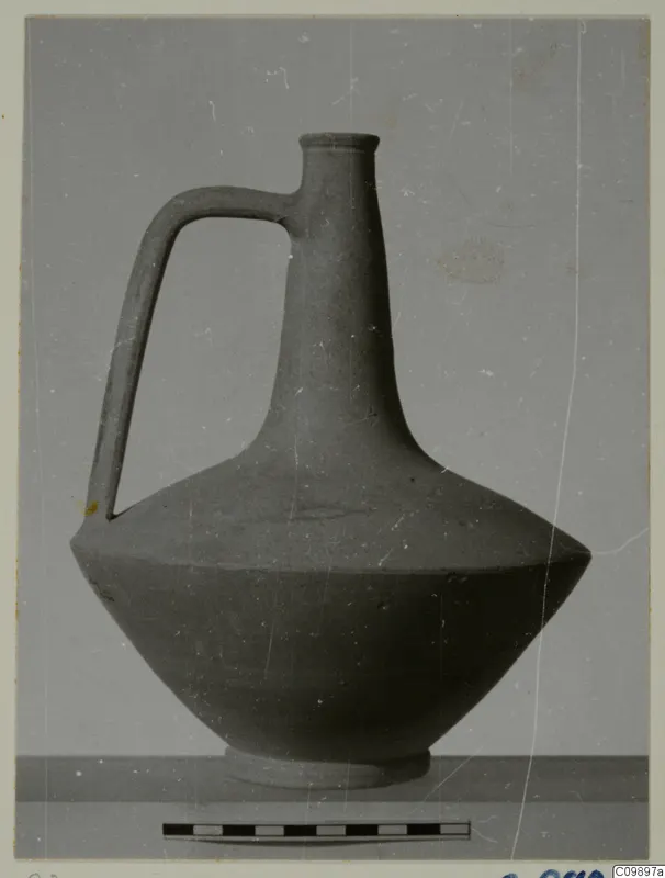 Jug with a tall neck and single loop handle, shown beside a scale bar against a plain grey backdrop