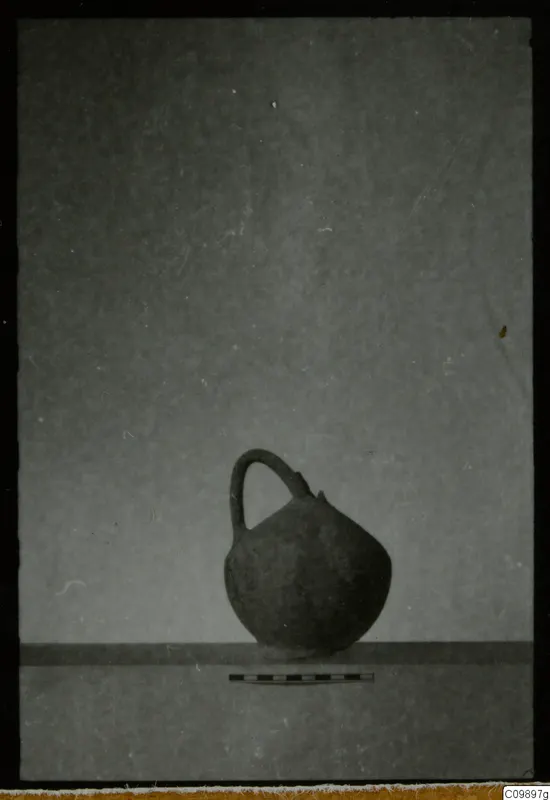 Pottery jug with a single loop handle and a scale bar in front, photographed against a plain grey background.
