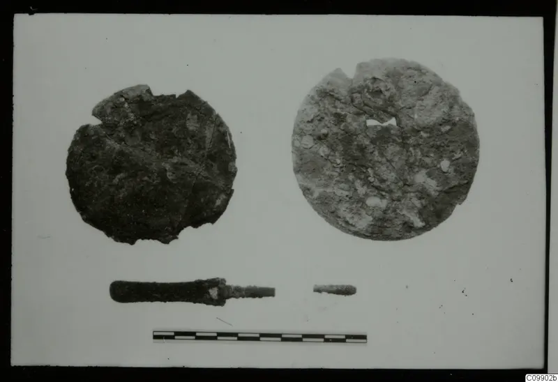 Two round, corroded discs, a rod-like tool and a small rod fragment on a white card, with a scale bar below.