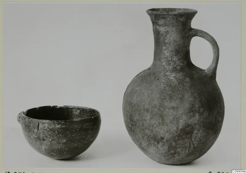 Dark grey pottery bowl and jug with a loop handle, standing side by side against a plain backdrop.