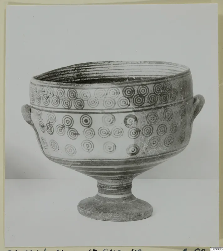 Footed bowl with two small handles and rows of concentric circles on its body, photographed against a plain backdrop