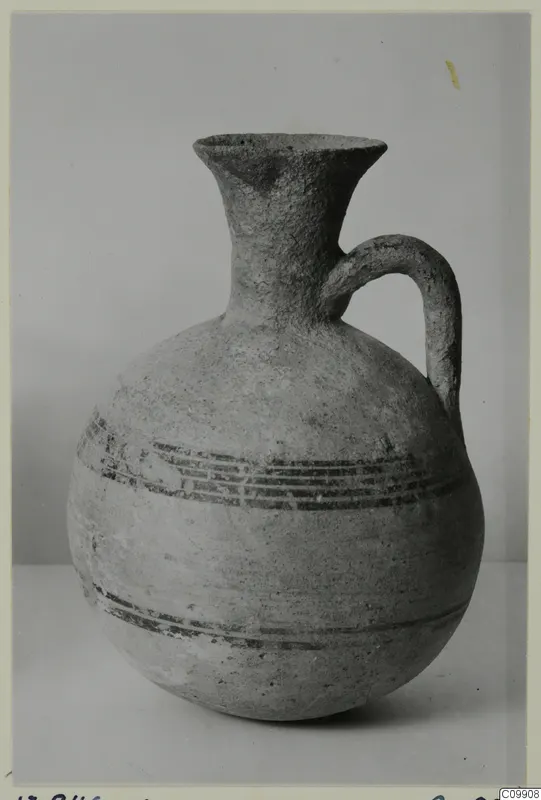 Pottery jug with a flaring neck, single strap handle and dark painted horizontal bands, photographed against a grey backdrop.