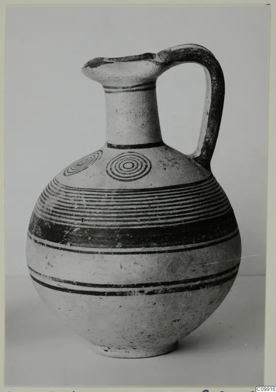 White Painted IV jug with a round body, tall neck and strap handle, decorated with dark bands and concentric circles