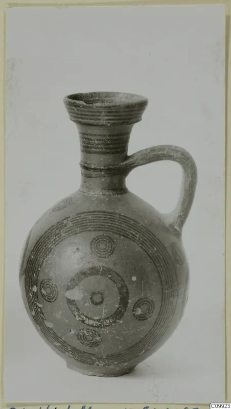 Round-bodied jug with a flaring mouth, a single strap handle and dark concentric circle painting, shown against a grey backdrop.