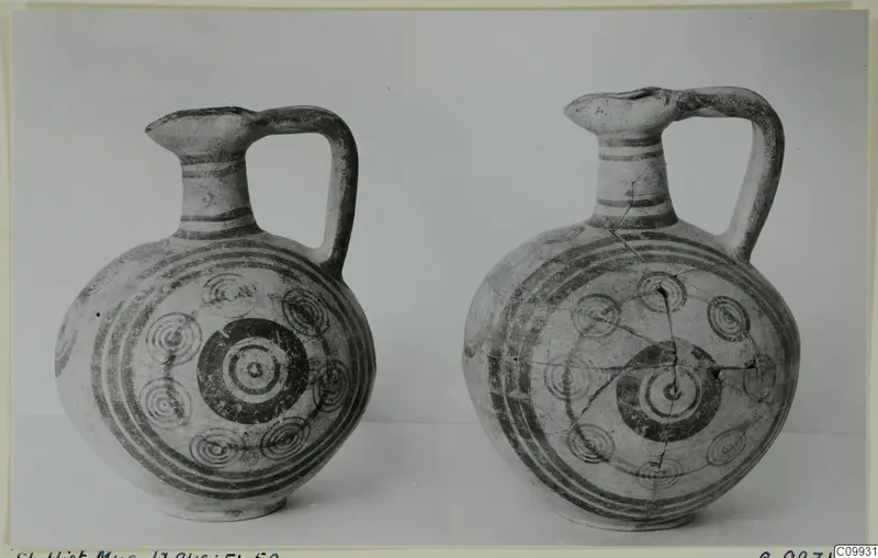Two painted pottery jugs with concentric-circle decoration and single handles, photographed side by side against a grey backdrop.