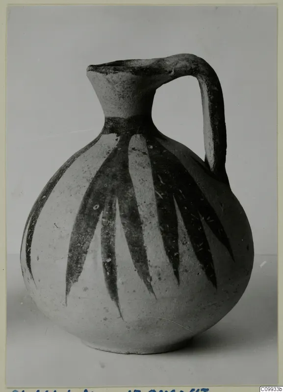 Round-bodied jug with a tall neck, strap handle and dark leaf-like painted strokes on a pale surface.