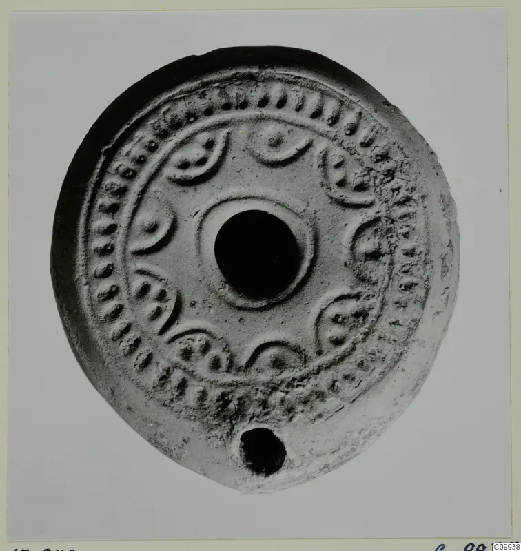 Round pale clay lamp seen from above, with a central filling hole and relief dots and crescent motifs around the rim