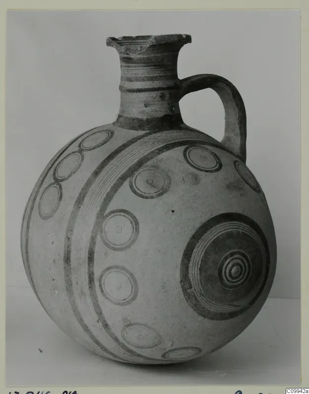 Round-bodied jug with one loop handle and dark painted concentric circles and bands on a pale surface.